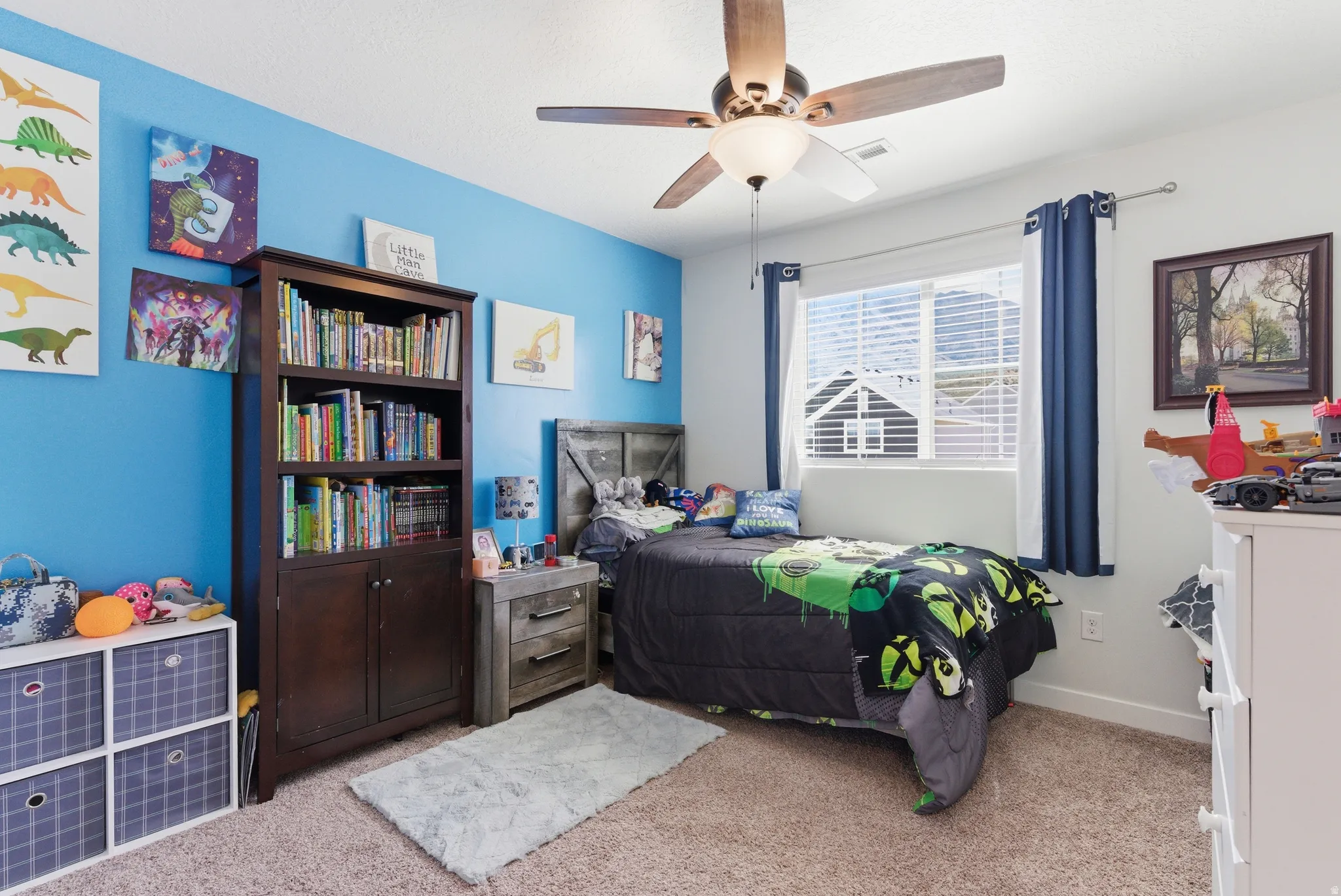 Ceiling fan with light fixture, single window with blinds, wood-finish headboard, built-in wooden bookcase, and carpeted flooring