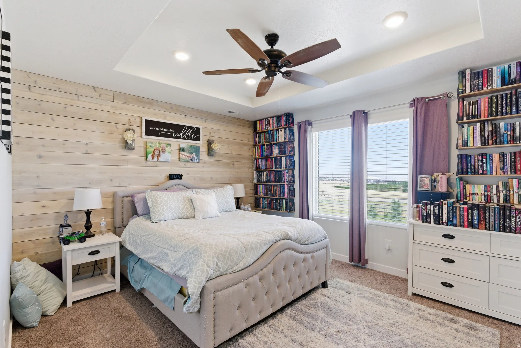 Tray ceiling with recessed lighting and a ceiling fan, accent wall with horizontal wood paneling, built-in shelving, and dual windows