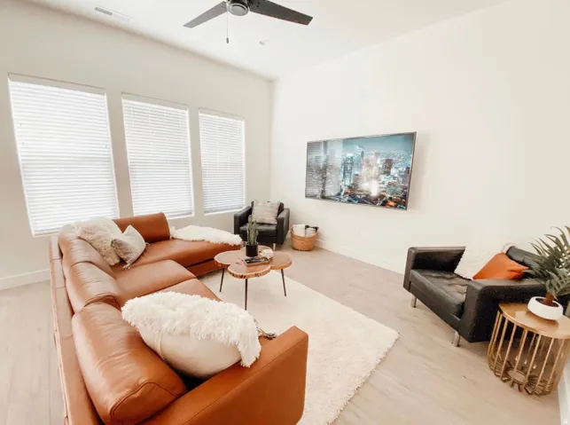White walls, wood-finish flooring, three large windows with blinds, a ceiling fan, and a wall-mounted display