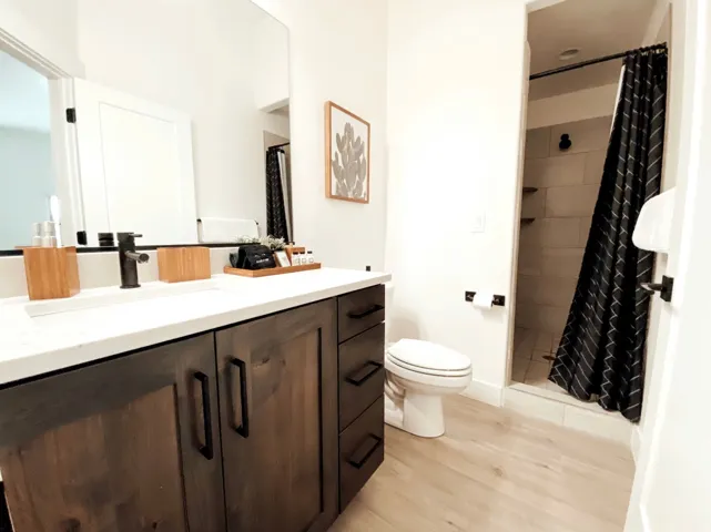 Wood-finish flooring, dark wood vanity with matte black hardware, white countertop with integrated sink, black wall-mounted faucet, and a tiled shower with recessed shelving
