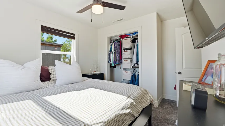 White walls, a ceiling fan with light, a window with dark blinds, a built-in closet with shelving, and gray carpeted flooring
