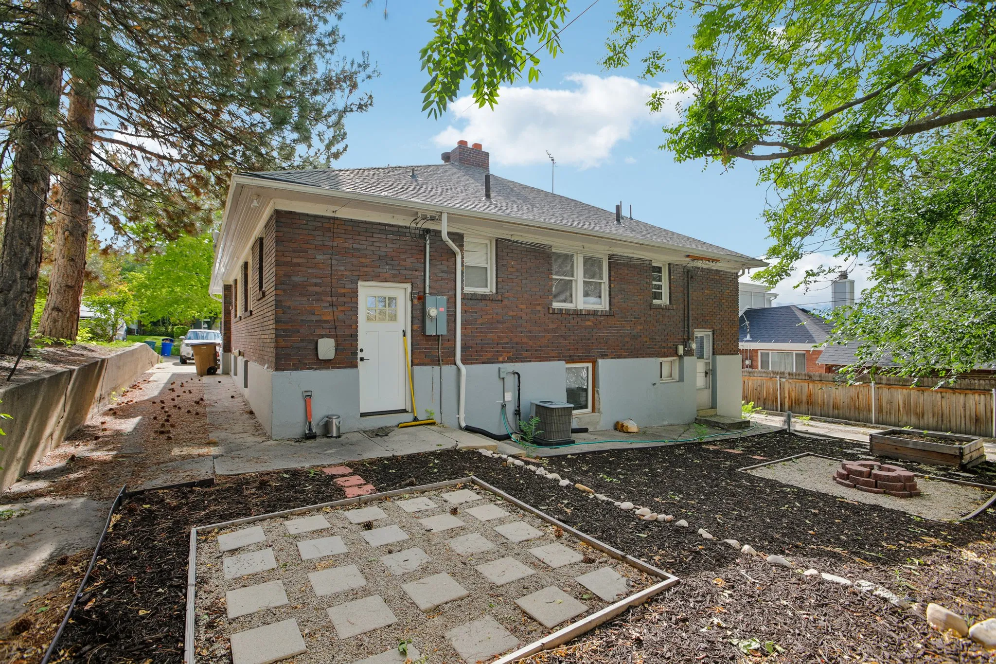 Brick exterior with a white door, framed by mature trees, features a concrete pathway and a paved patio area