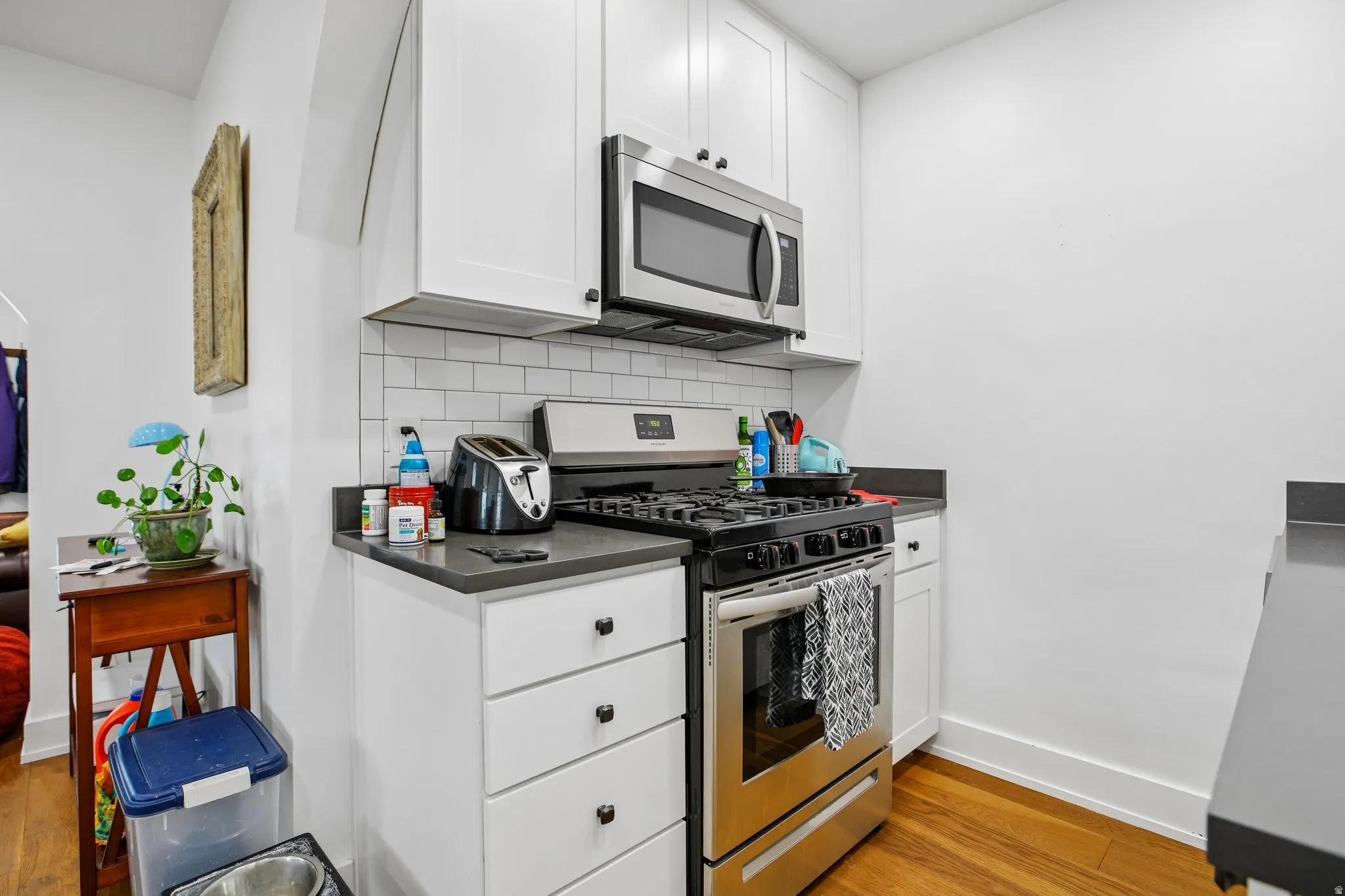 White shaker cabinetry, stainless steel appliances, subway tile backsplash, dark countertops, and wood-finish flooring