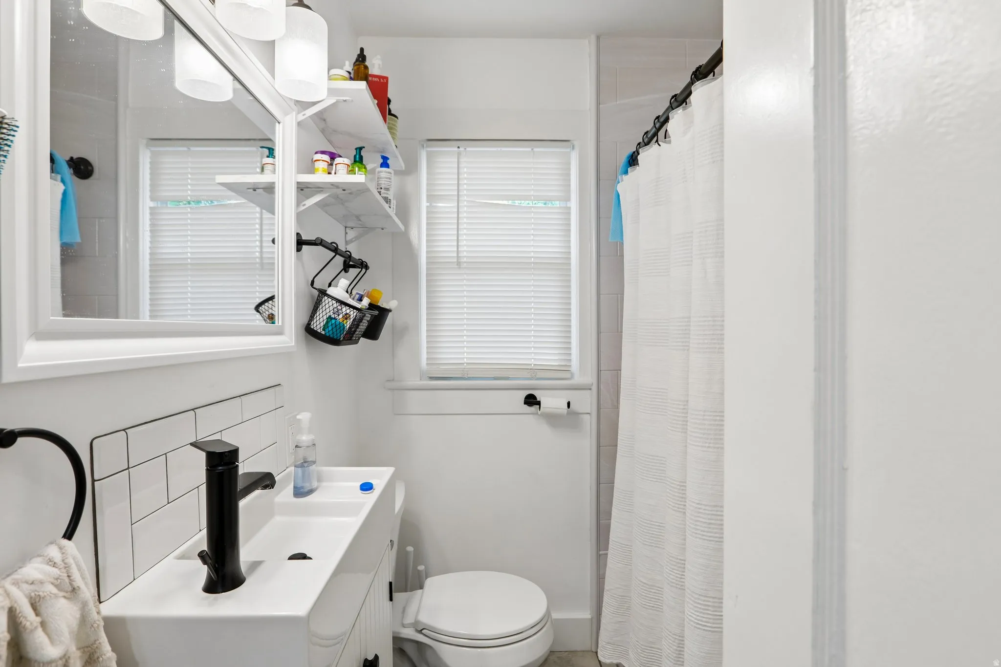 White subway tile backsplash, contemporary black faucet, white rectangular vessel sink, white vanity with black hardware, and an overhead vanity light fixture