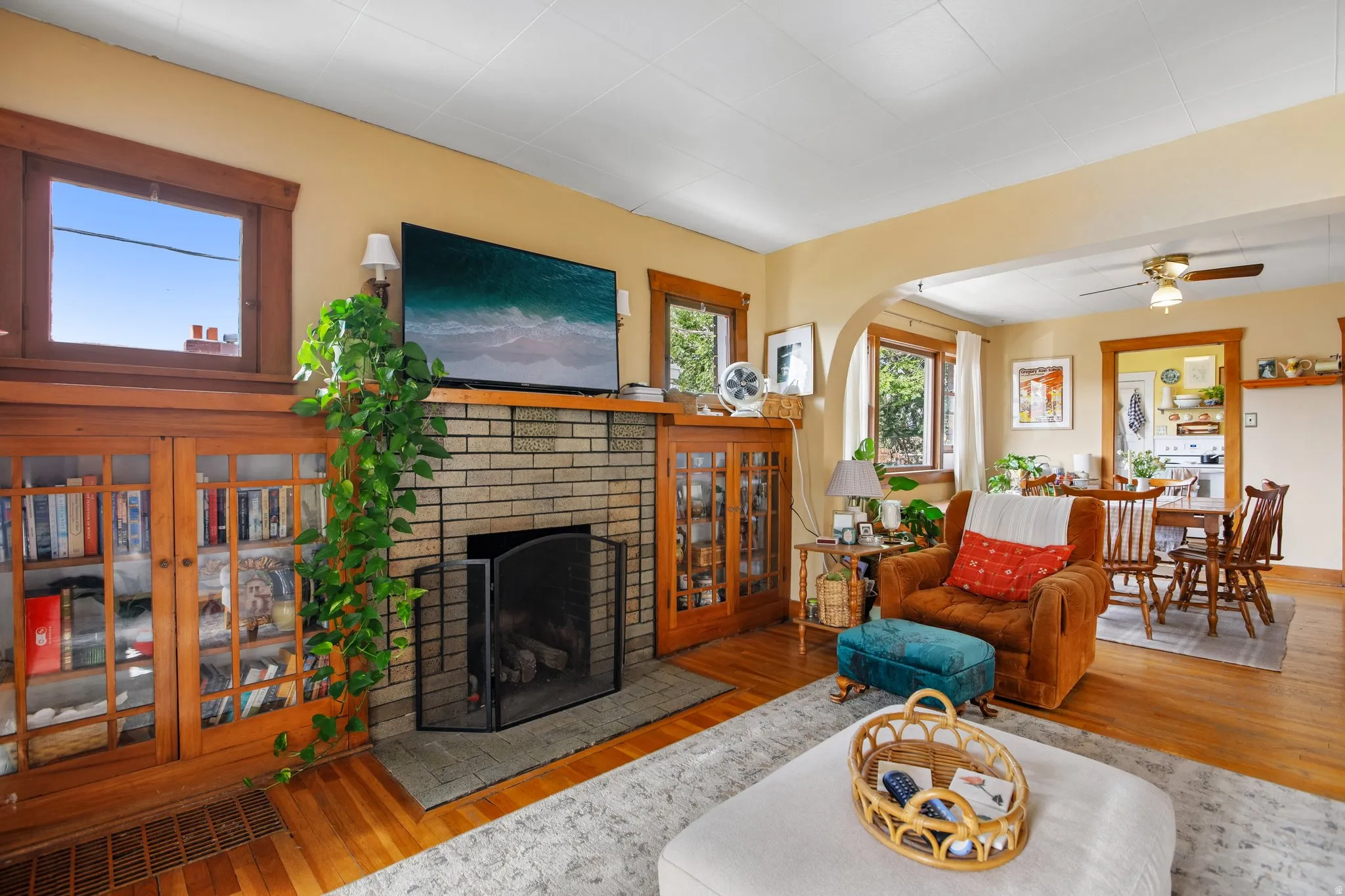 Hardwood flooring, brick fireplace with mantel, built-in cabinetry with glass doors, cased windows, and an arched entryway
