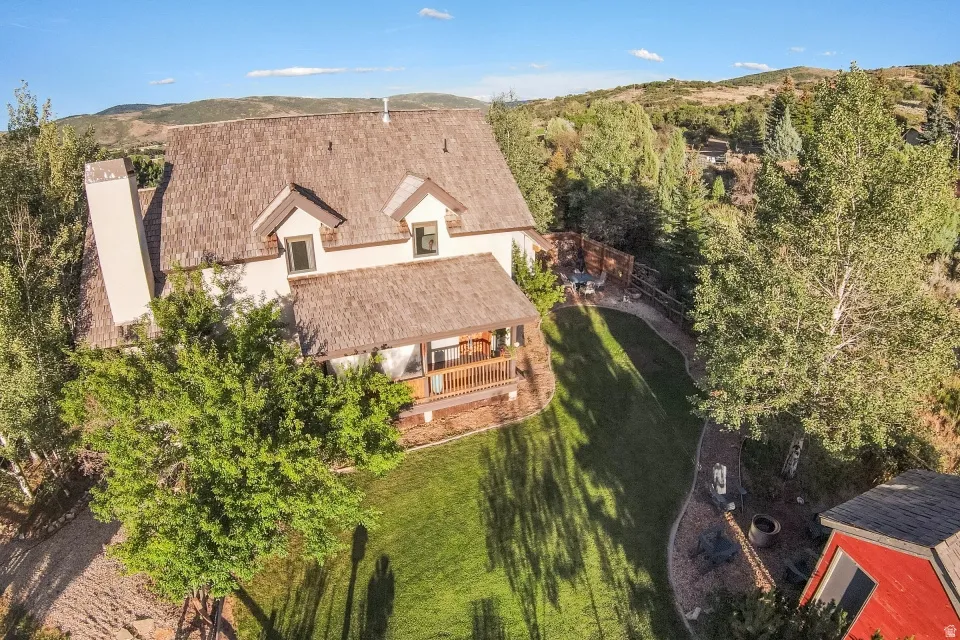 Gable roof structure with dormer windows, covered porch, exterior chimney, extensive lawn, and mature trees