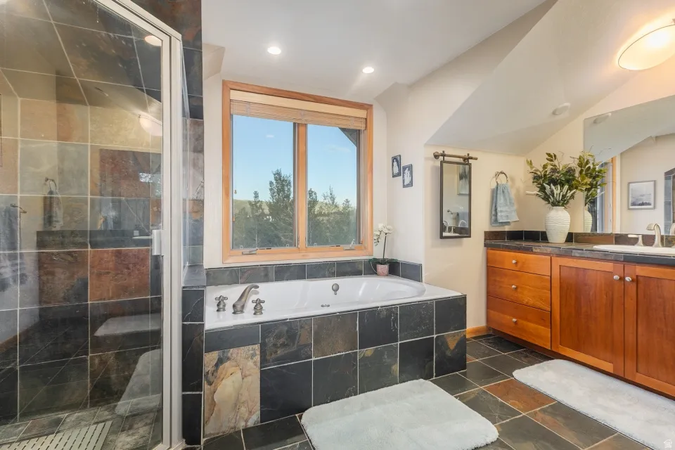 Tiled flooring and bath surround, built-in soaking tub, glass-enclosed shower, wood-finish vanity with countertop, and a window with wood trim
