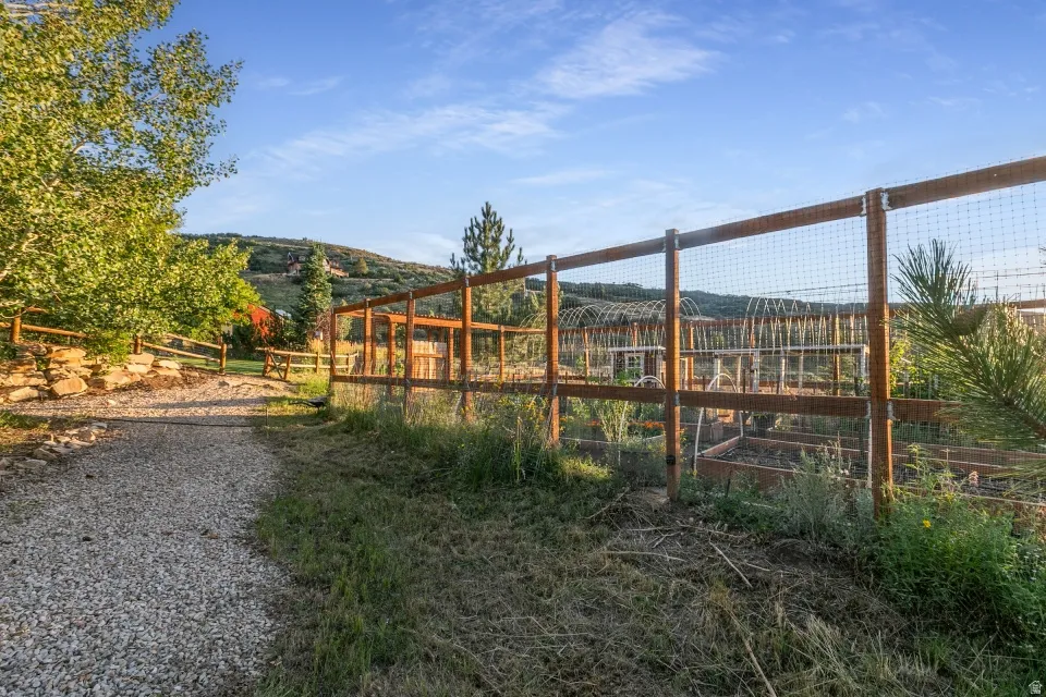 Gravel pathway leading to a fenced garden with raised beds and trellises, bordered by natural landscaping and mature trees