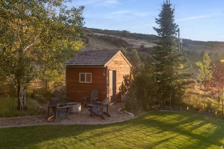 Manicured lawn with a gravel patio featuring a fire pit, bordered by mature trees and a rustic wood-sided shed with an asphalt shingle roof