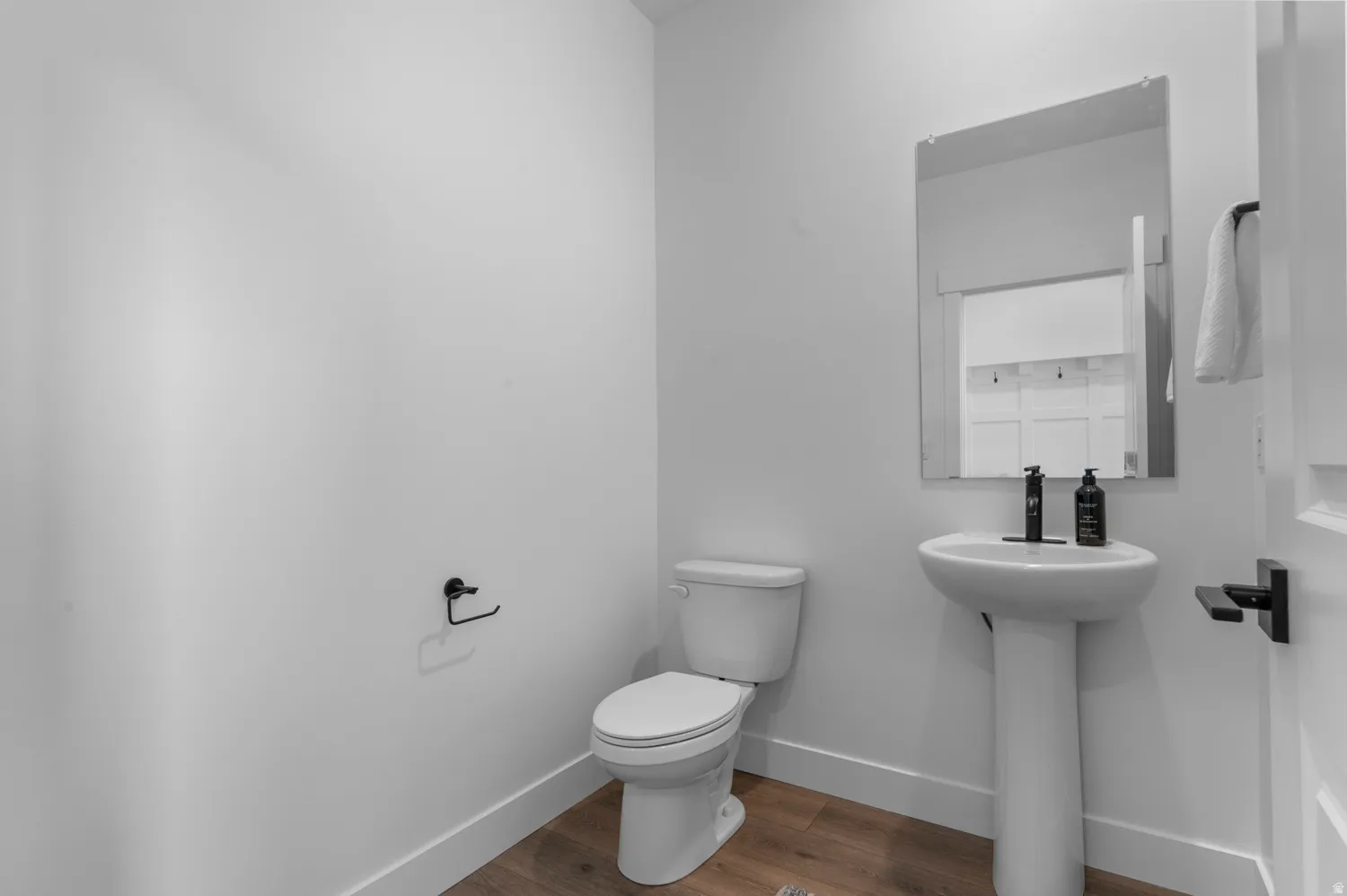 Pedestal sink with black faucet, frameless mirror, toilet, wood-finish flooring, and white trim