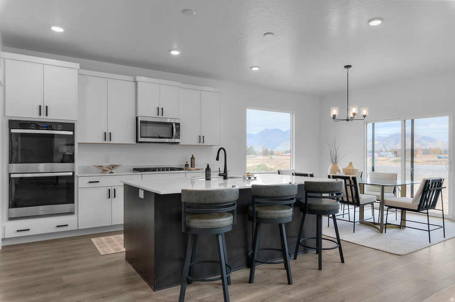 Kitchen featuring white cabinetry, stainless steel double wall ovens, a central island with a light-toned countertop, wood-finish flooring, and a dining area with a contemporary chandelier