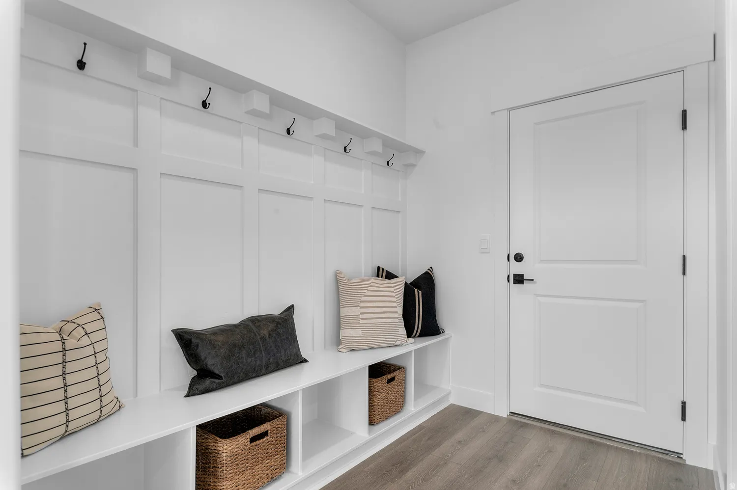 Built-in storage bench with cubbies, board and batten wall paneling, integrated coat hooks, wood-finish flooring, and a paneled interior door with matte black hardware