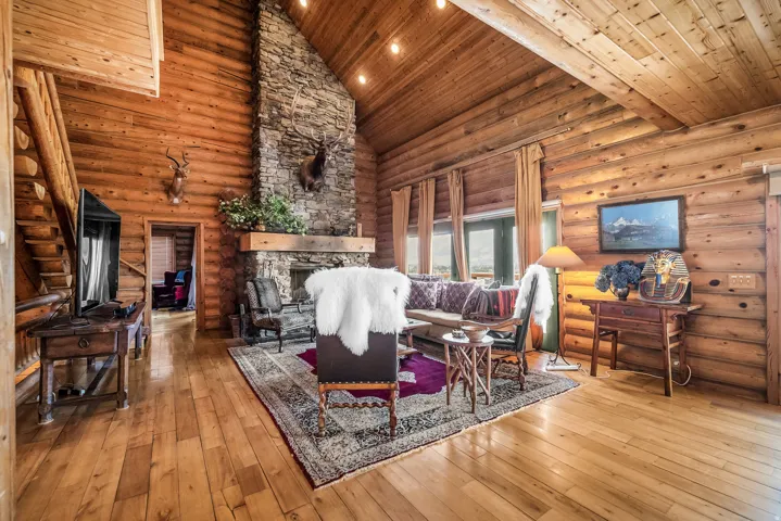 Stone fireplace with a wood-beam mantel, exposed log walls, wood-finish flooring, vaulted wood ceiling, and a wood staircase with a log railing