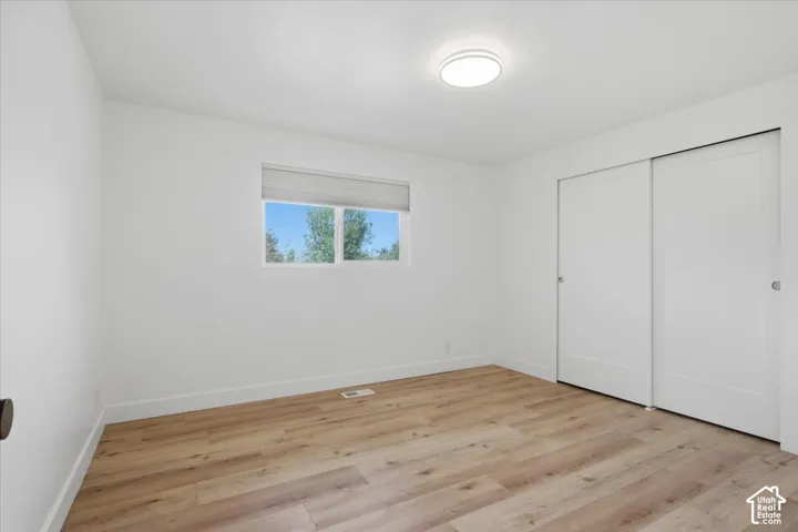 Another bedroom with bright room featuring wood-finish flooring, a flush-mount ceiling light, a window with a roller shade, and sliding closet doors