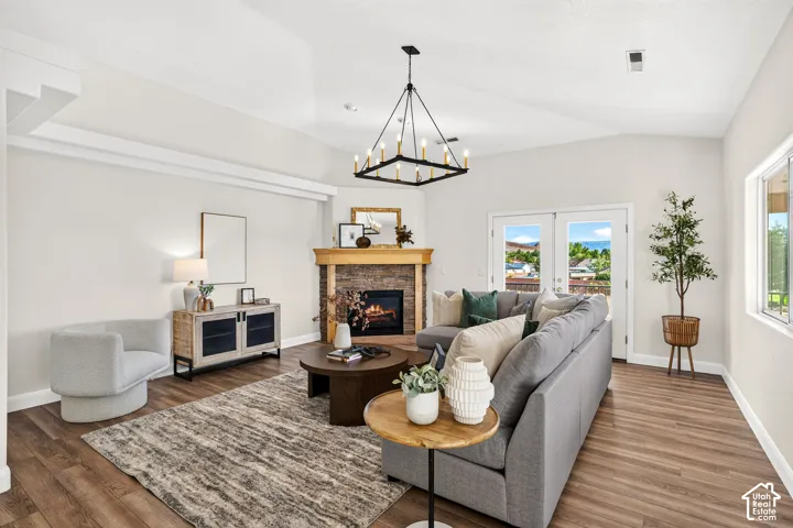 Vaulted ceiling with an angular chandelier, stone-faced fireplace with a wood mantle, wood-finish flooring, and white double French doors