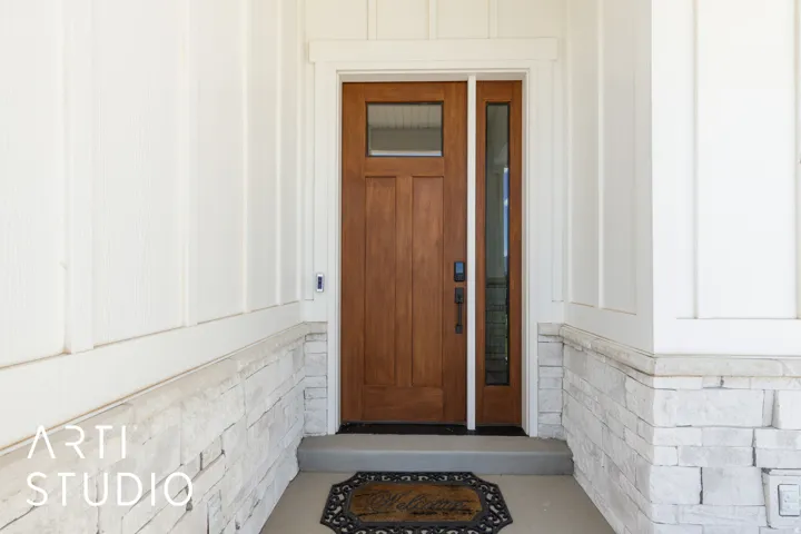 Solid wood entry door with side light and transom window, framed by white trim and board and batten siding, above a stone veneer foundation