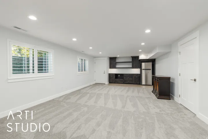 Recessed lighting, light gray carpeting, two windows with white plantation shutters, built-in dark wood cabinetry with open shelving, and a stainless steel refrigerator