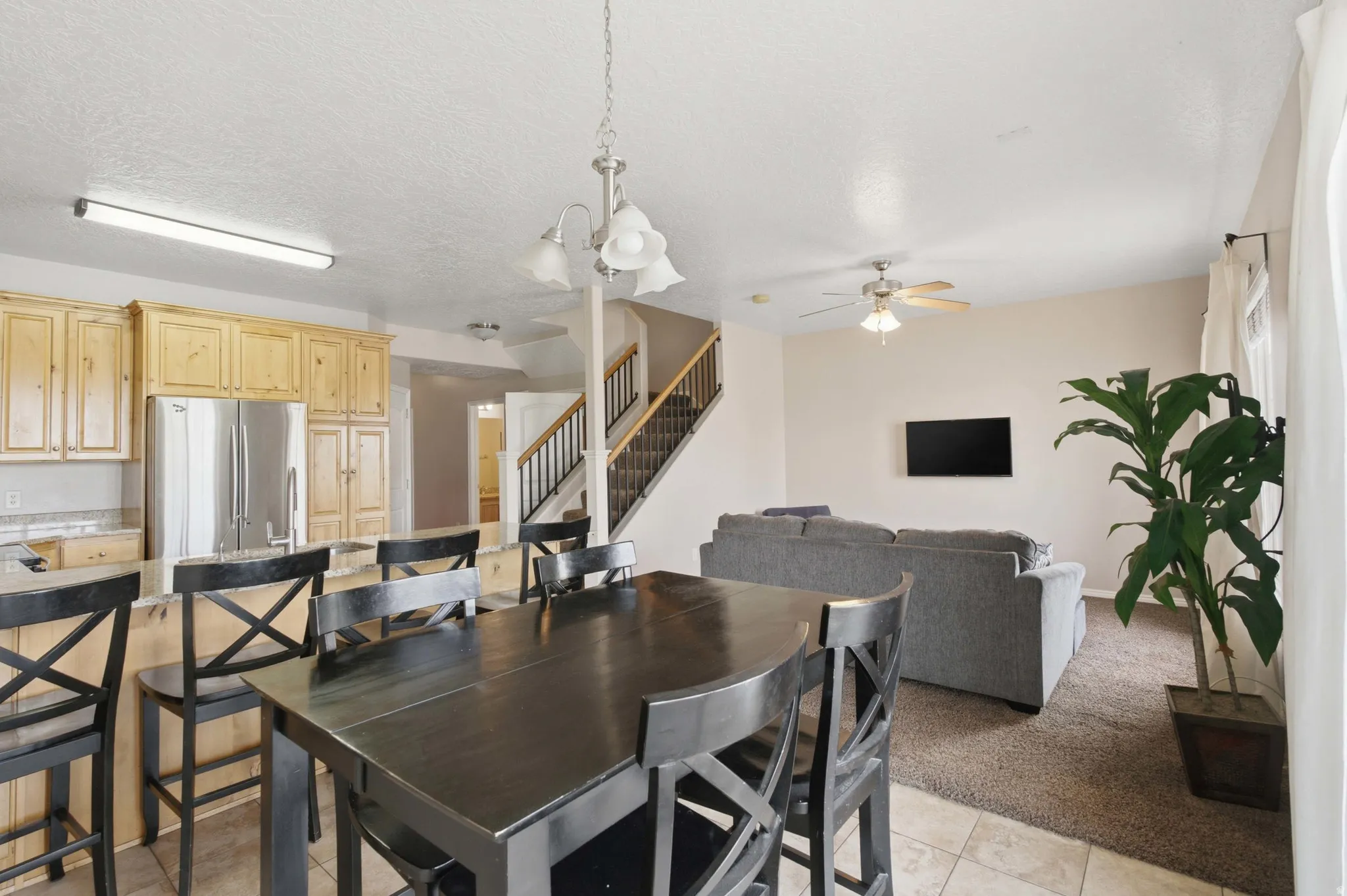 Open concept living area featuring a kitchen with light wood cabinetry, stainless steel refrigerator, and a breakfast bar, alongside a carpeted living space with a ceiling fan, and a staircase with black railings