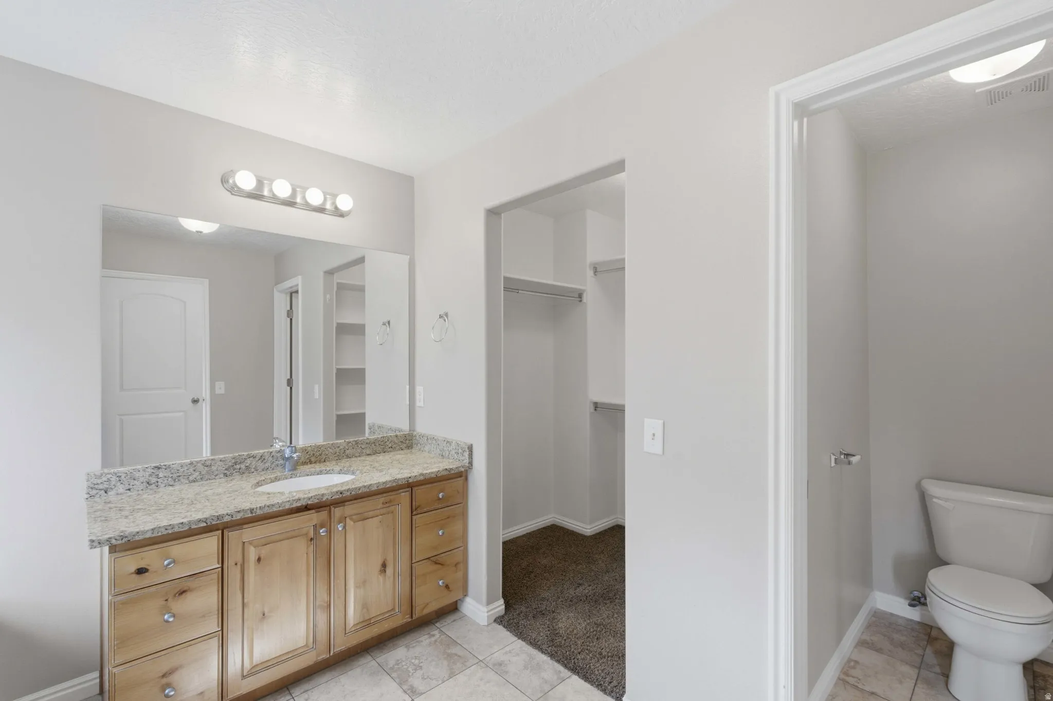Master bathroom suite featuring dual vanity with wood cabinetry and granite countertop, large framed mirror with integrated lighting, ceramic tile flooring, and direct access to walk-in closet and toilet room