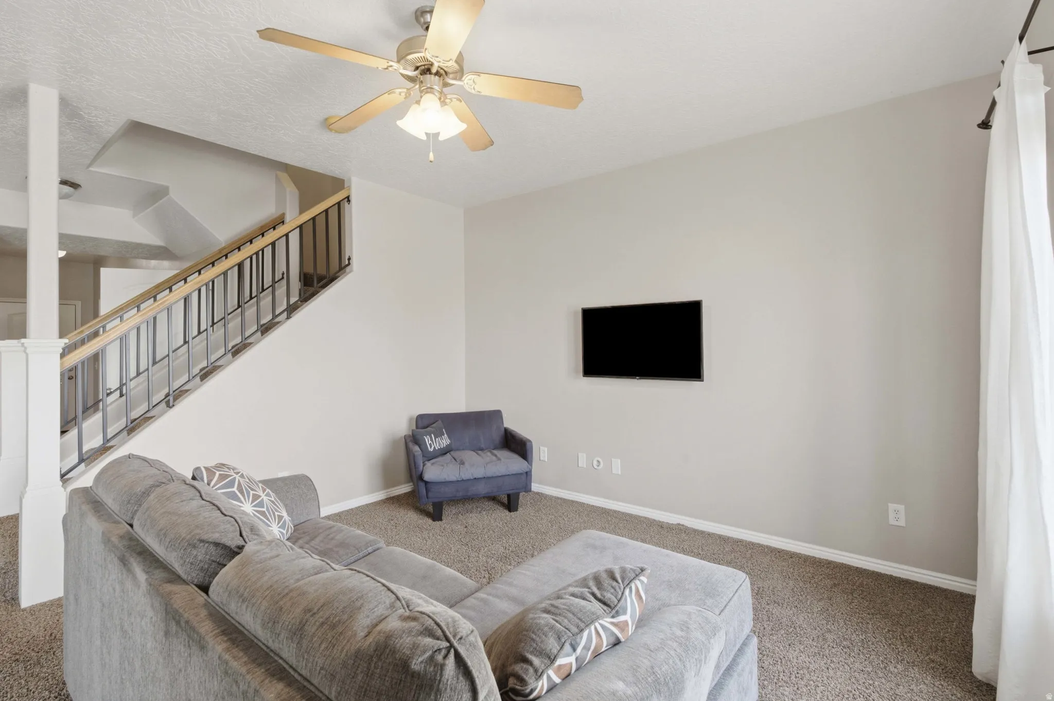 Spacious lower-level recreation room featuring neutral carpet flooring, a ceiling fan with integrated lighting, a flat-screen television mount, and abundant natural light from large windows