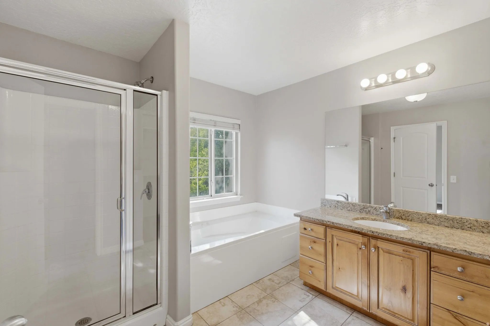 Spa-inspired primary bathroom featuring a soaking tub with natural light from a casement window, dual-sink vanity with wood cabinetry and stone countertops, frameless glass shower enclosure, tile flooring, and recessed and vanity lighting