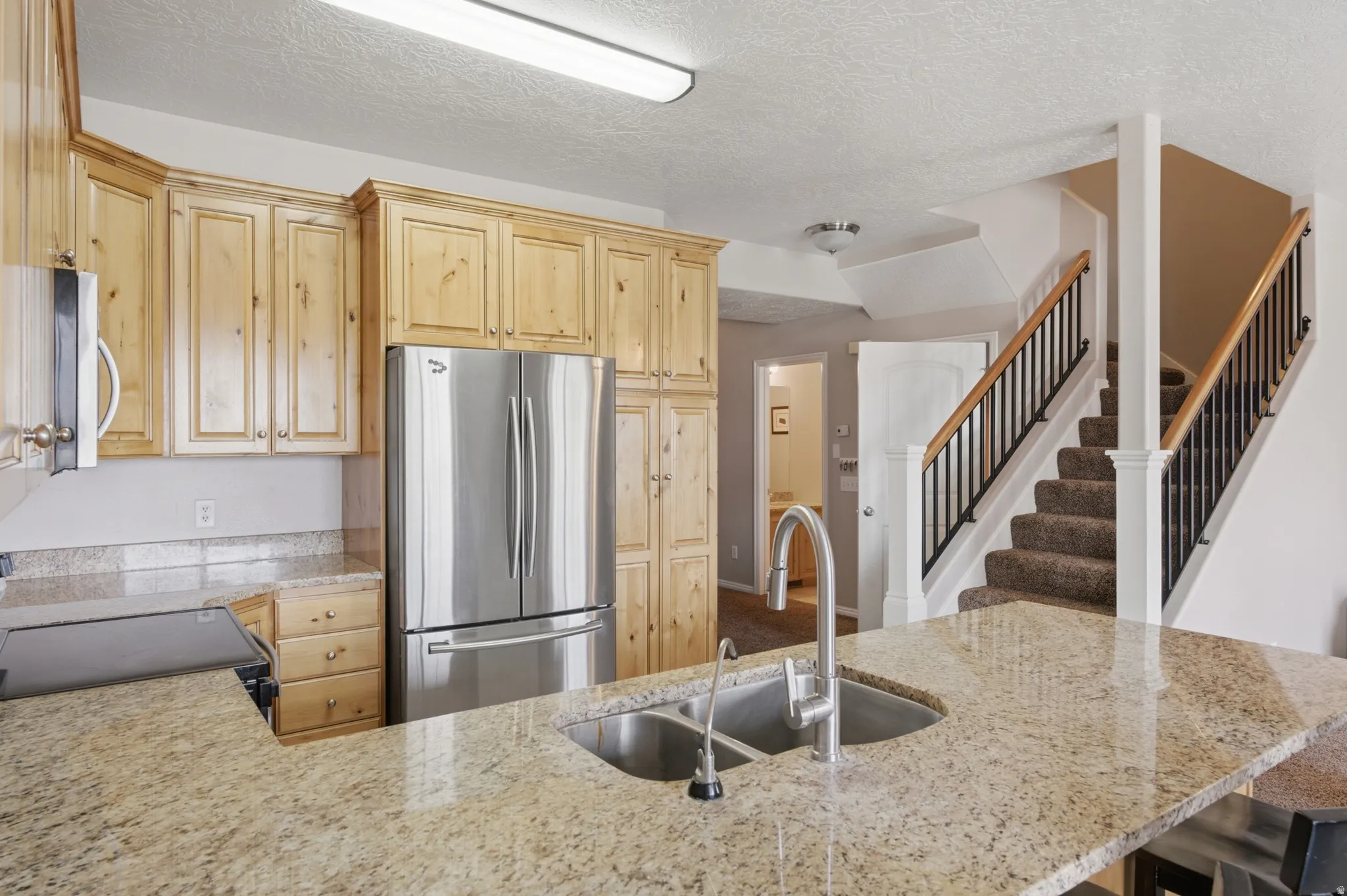 Granite countertops, stainless steel appliances, wood cabinetry, stainless steel double basin sink, and a carpeted staircase with wood handrails