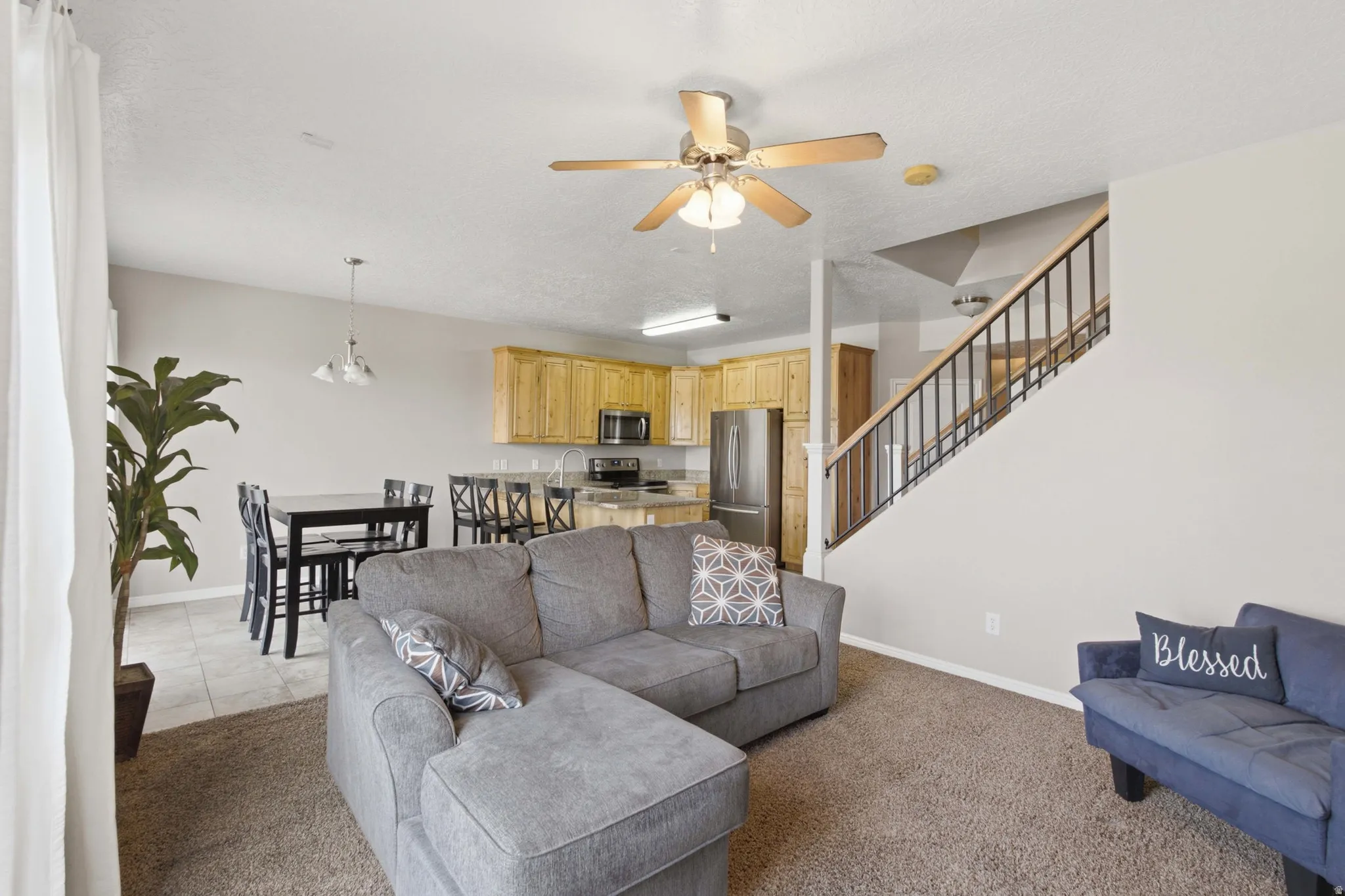 Open-concept living area featuring a gray upholstered sectional sofa with chaise lounge, neutral carpet flooring, and a wood-finish ceiling fan with integrated lighting