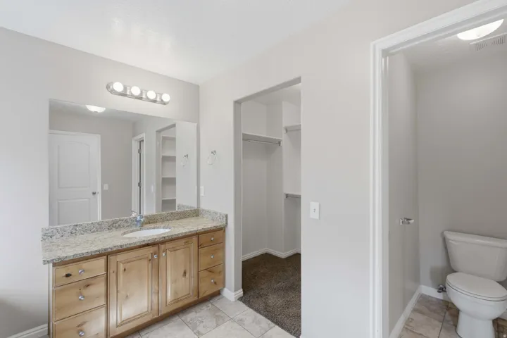 Master bathroom suite featuring dual vanity with wood cabinetry and granite countertop, large framed mirror with integrated lighting, ceramic tile flooring, and direct access to walk-in closet and toilet room