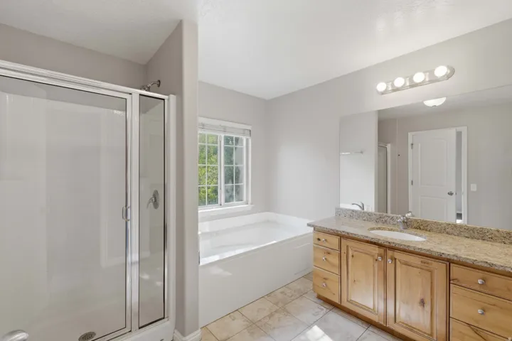 Spa-inspired primary bathroom featuring a soaking tub with natural light from a casement window, dual-sink vanity with wood cabinetry and stone countertops, frameless glass shower enclosure, tile flooring, and recessed and vanity lighting
