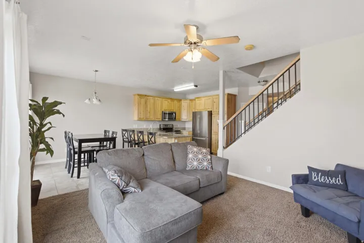 Open-concept living area featuring a gray upholstered sectional sofa with chaise lounge, neutral carpet flooring, and a wood-finish ceiling fan with integrated lighting
