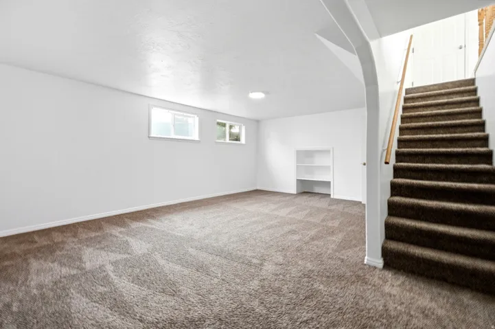 Carpeted flooring, white painted walls, built-in shelving, two windows, and a carpeted staircase with a wood handrail