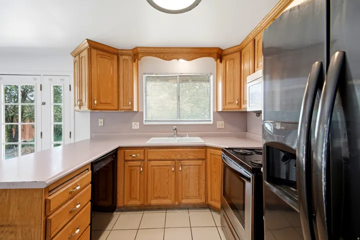 Wood cabinetry, light-toned countertops, white tile flooring, stainless steel appliances, and French patio doors