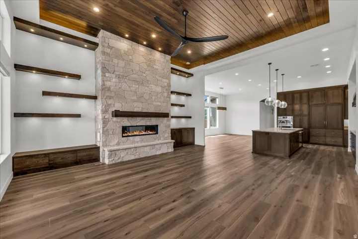 Wood-finish flooring throughout, a stone-clad fireplace, integrated floating shelves with accent lighting, a wood-paneled ceiling, and a central kitchen island with pendant lighting