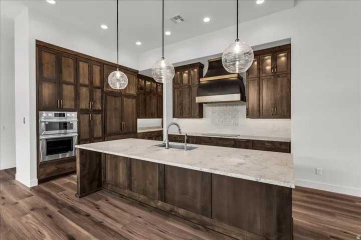 Kitchen featuring custom wood cabinetry, stainless steel double ovens, a large island with a light-toned countertop, and wood-finish flooring