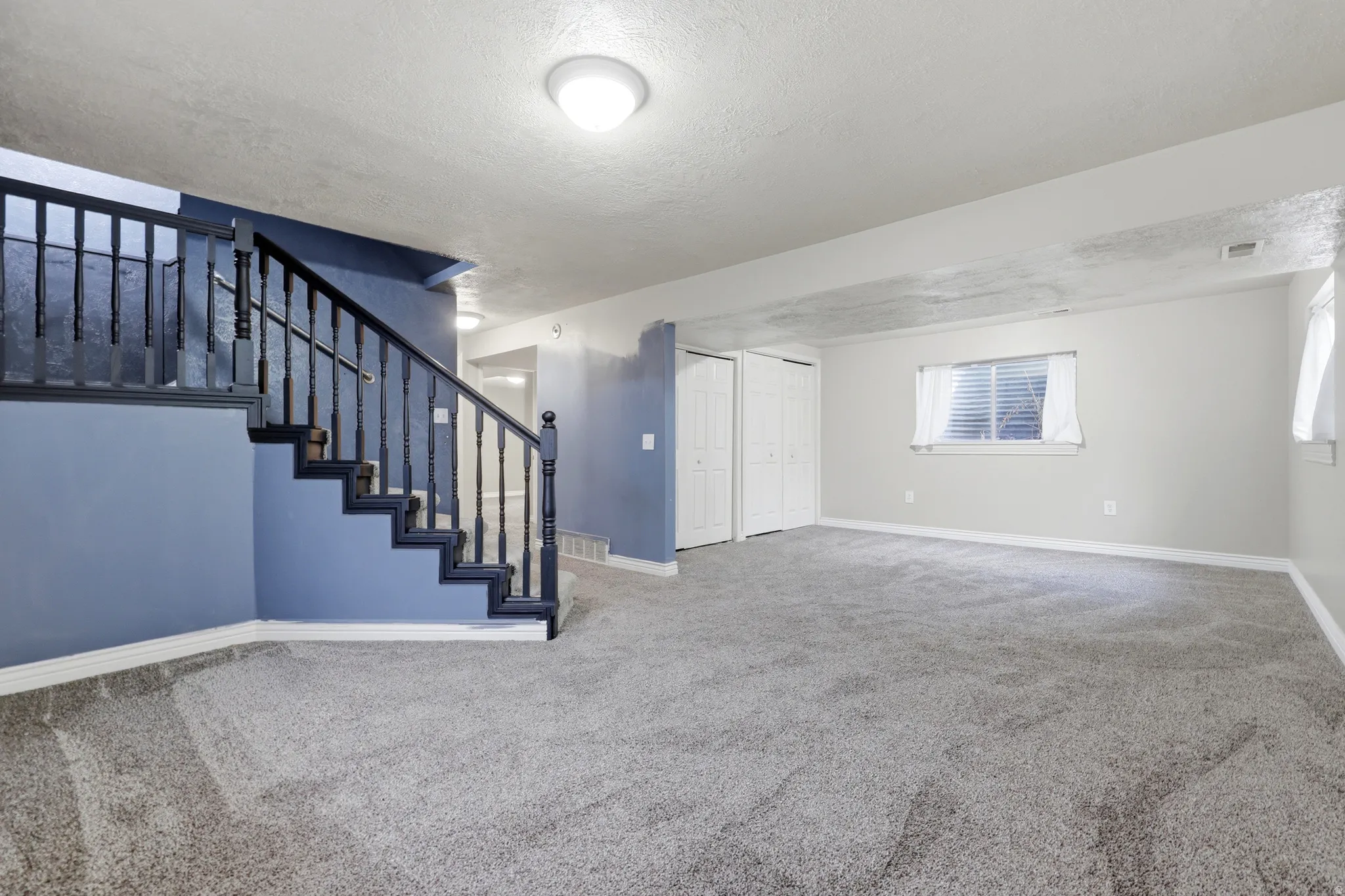 Spacious basement recreation area featuring neutral carpeting throughout, recessed ceiling lighting, and a custom staircase with black metal railings and blue-painted walls