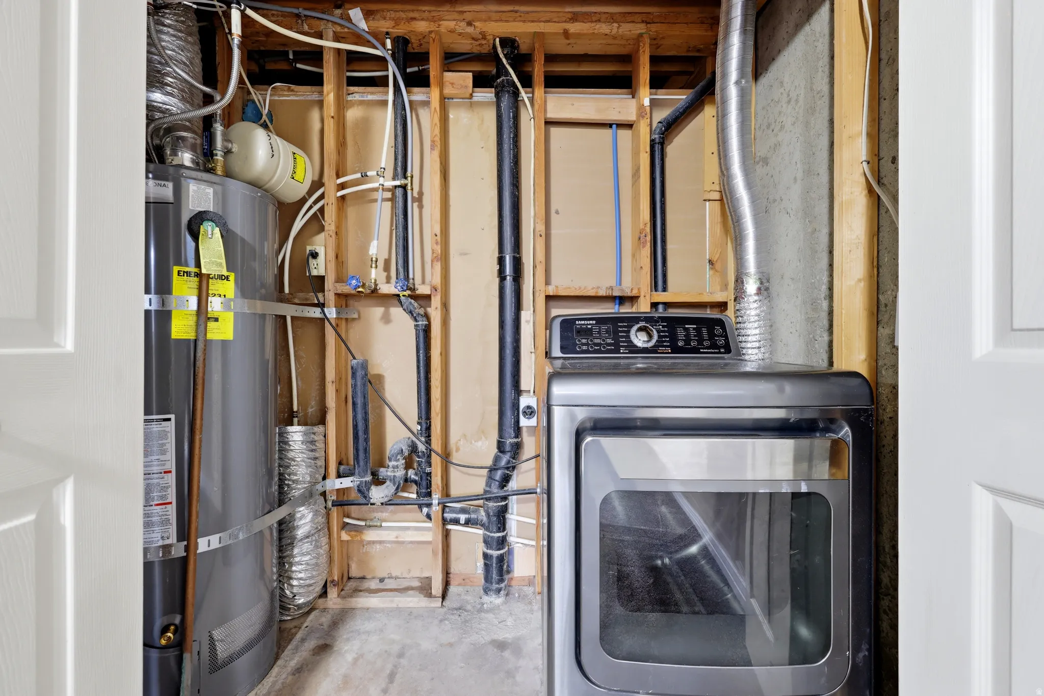 Utility room featuring modern front-load dryer with digital controls, water heater with insulation wrap, flexible supply lines, and wooden framed structural elements