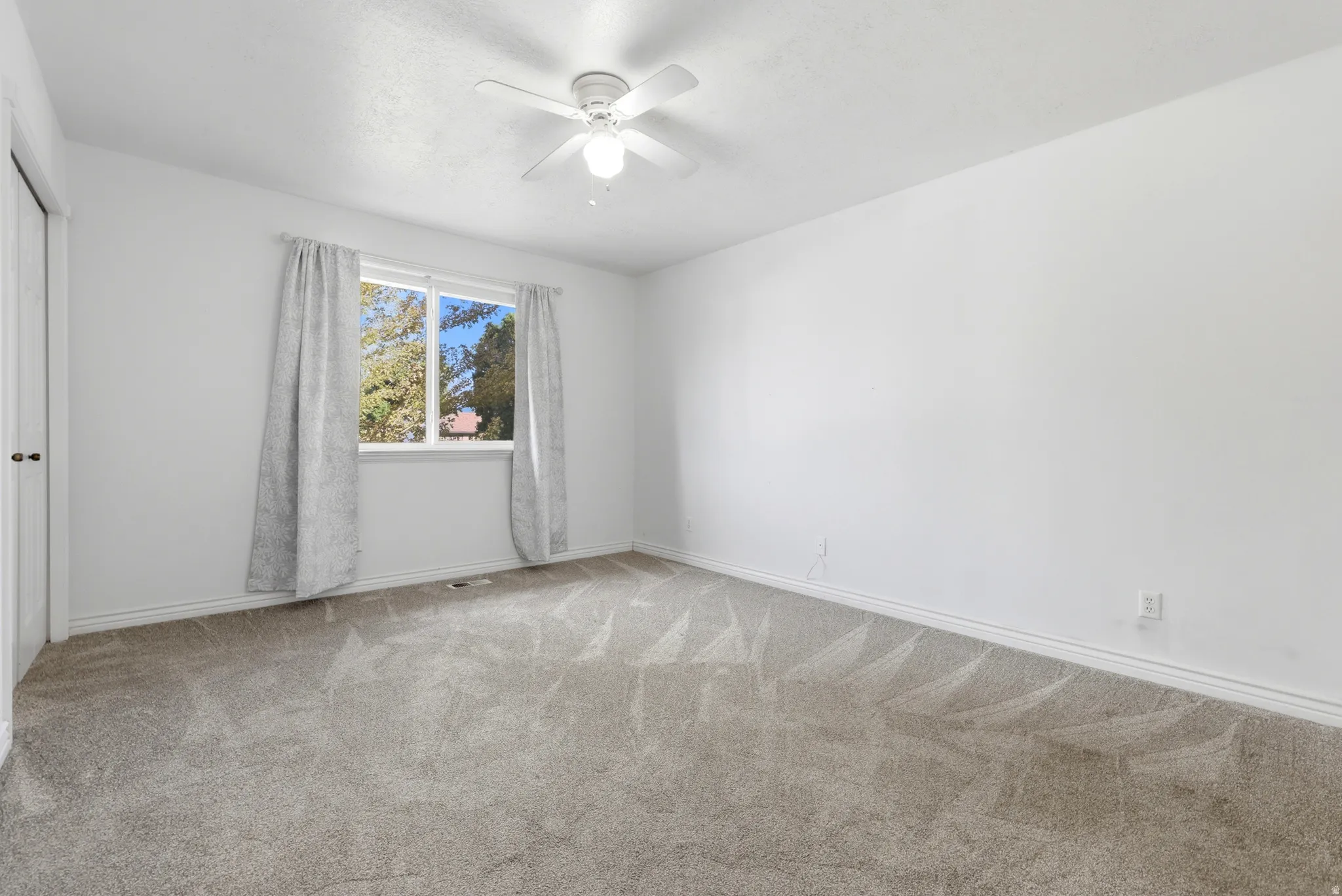 Neutral wall paint, ceiling fan with integrated lighting, window with a natural backdrop, and gray carpet flooring