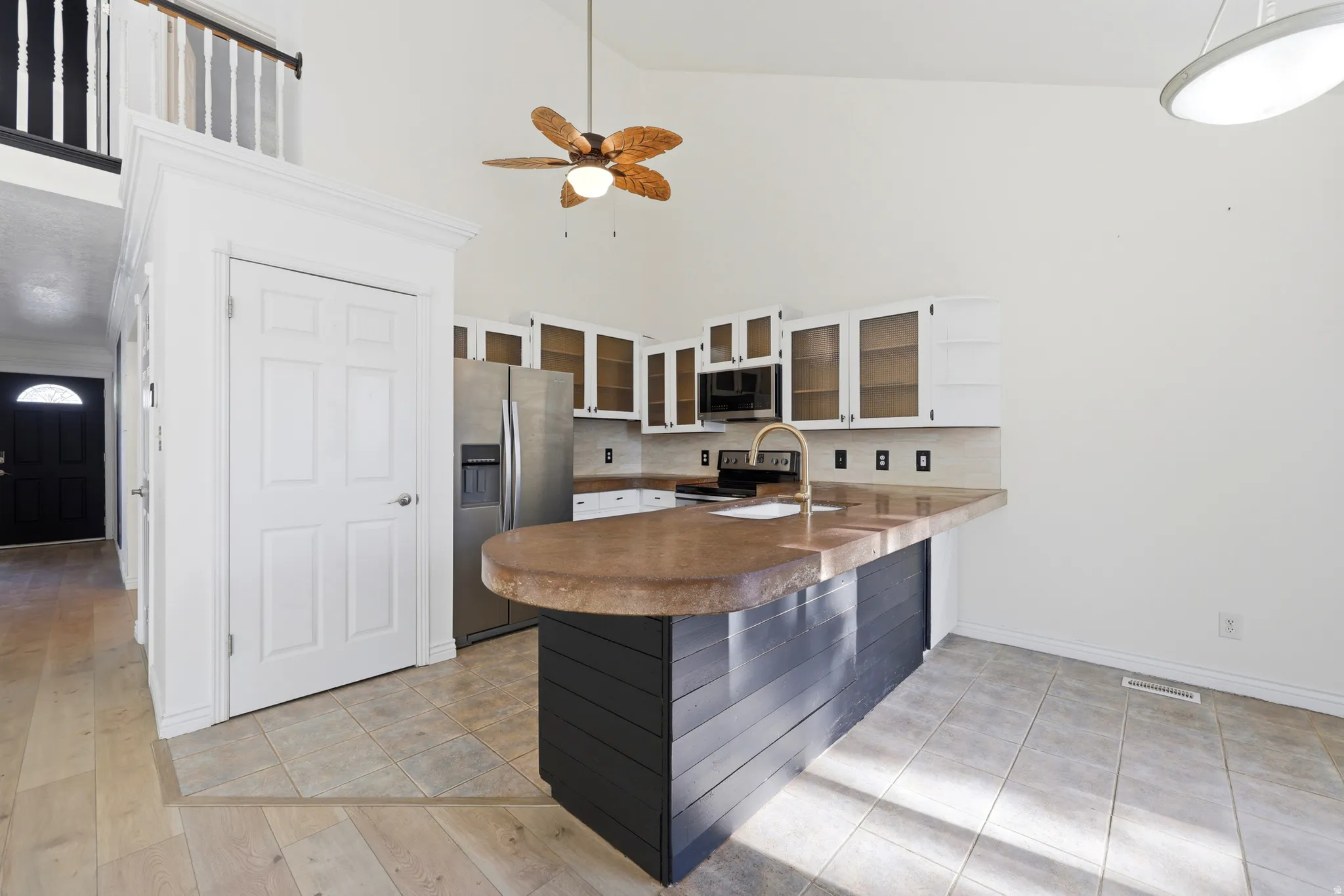 Open-concept kitchen featuring a curved wood-finish island with stainless steel base, granite countertops, stainless steel appliances, upper cabinetry with glass-front doors, tile flooring, ceiling fan, and abundant natural light from recessed and pendant