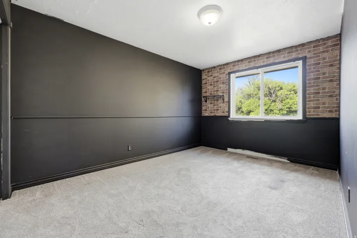 Spacious room featuring neutral carpeting, black accent wall paneling, exposed brick detailing, and a large window with natural light and outdoor scenery