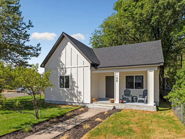 White board and batten siding, dark shingle roof, covered front porch, concrete steps, and mature perimeter trees