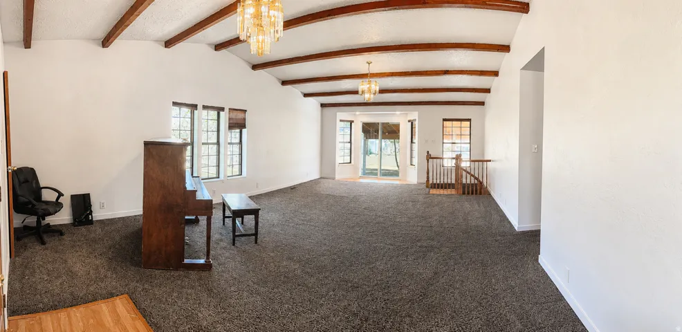 Vaulted ceiling with exposed wood beams, dual decorative chandeliers, multiple windows with blinds, and sliding glass doors