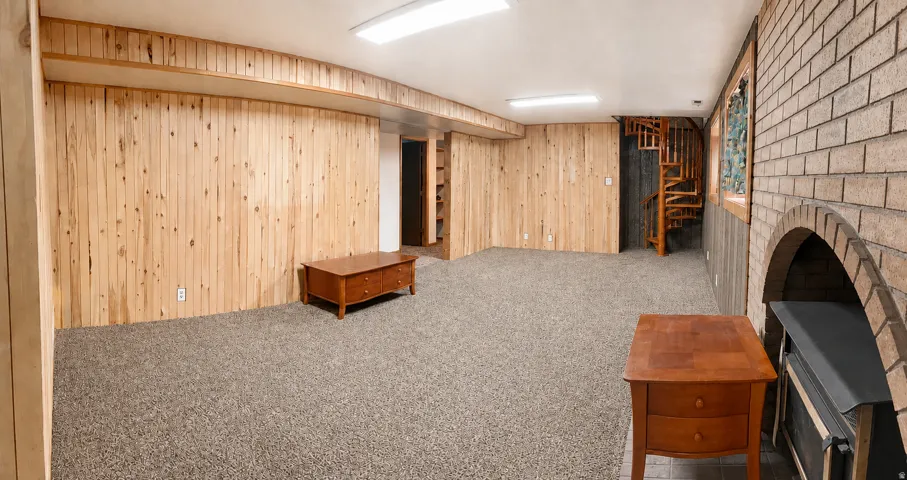 Spacious room featuring wood paneling, a brick fireplace with an arched opening, a spiral wooden staircase, and textured carpeting