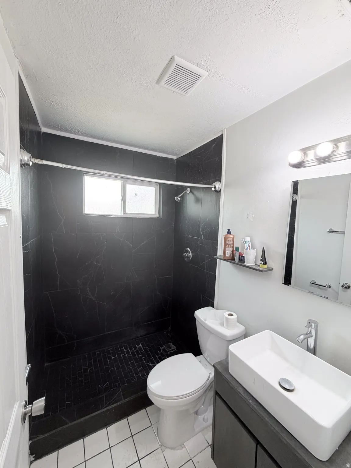 Modern bathroom featuring a walk-in shower with black tile surround and frameless glass enclosure, white subway tile flooring, white vessel sink with chrome faucet, floating vanity with dark finish, and large mirror with integrated lighting