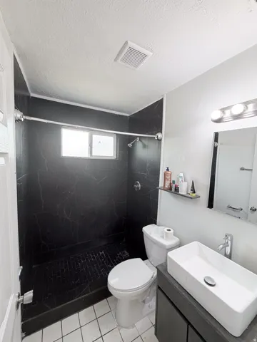 Modern bathroom featuring a walk-in shower with black tile surround and frameless glass enclosure, white subway tile flooring, white vessel sink with chrome faucet, floating vanity with dark finish, and large mirror with integrated lighting