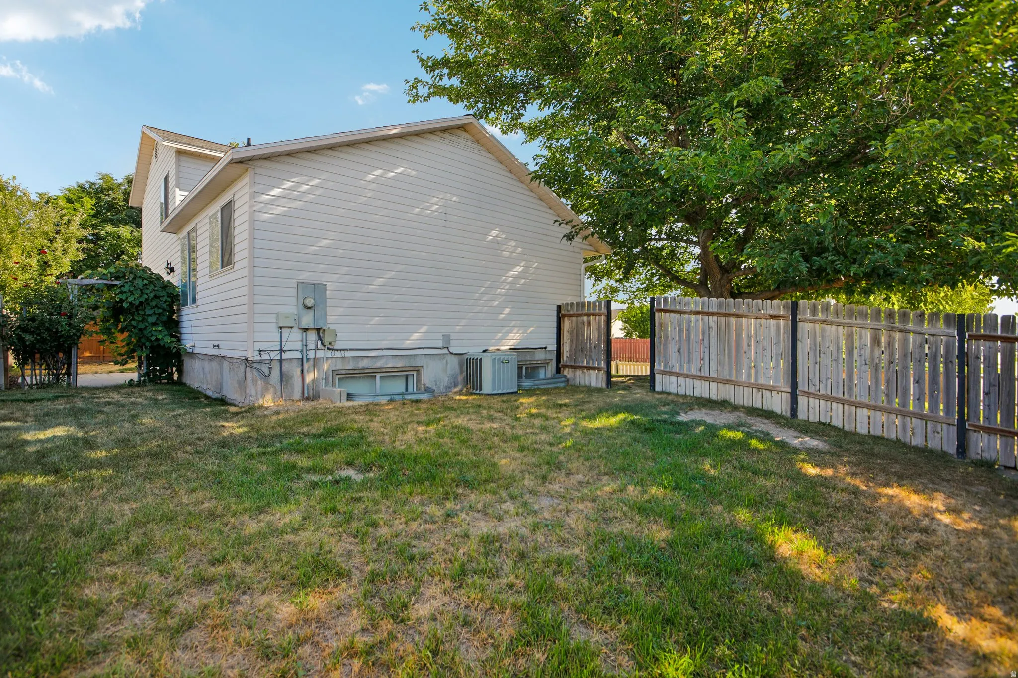 Exterior featuring white siding, a full fence, mature trees, and a ground-level window