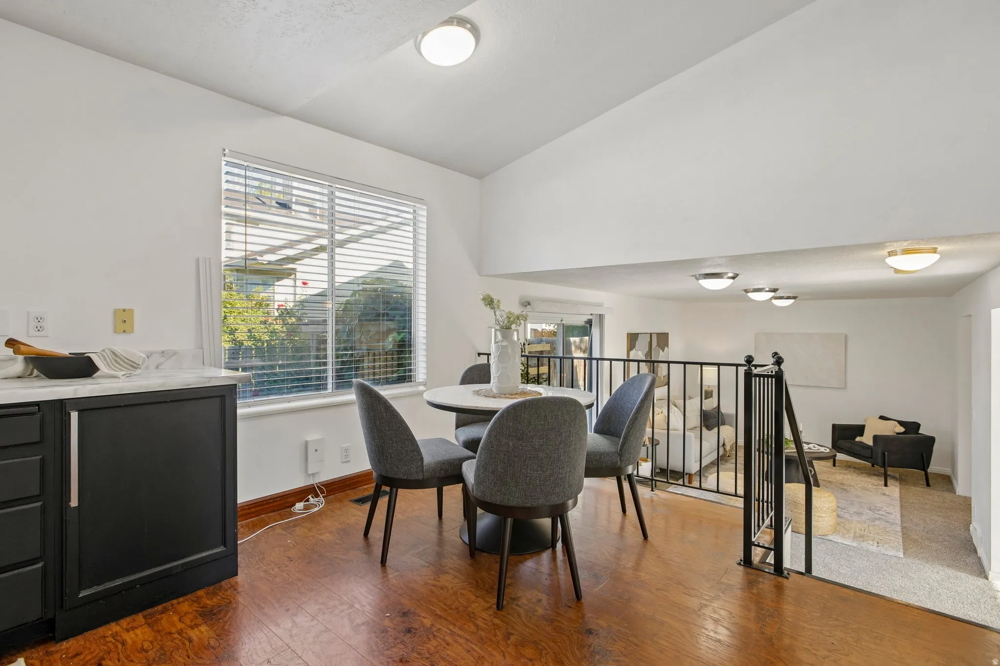 Wood-finish flooring, white walls, black cabinetry with light-toned countertops, large window with blinds, and a black metal railing
