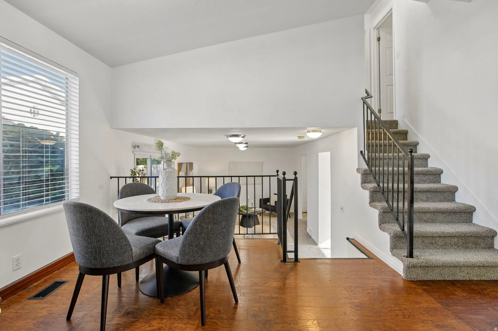 Wood-finish flooring, white walls, carpeted staircase with metal railings, and a window with blinds