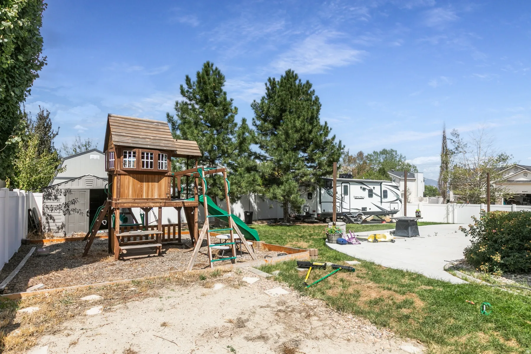 Expansive backyard featuring a wood play structure, green slide, white privacy fence, and mature pine trees