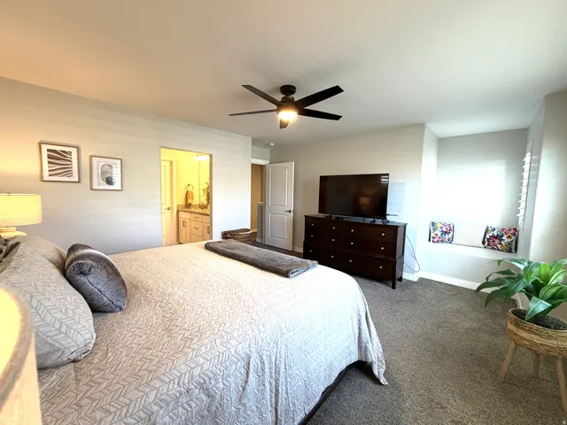 Recessed window seating, textured carpeting, ceiling fan with integrated lighting, and a doorway opening to a bathroom featuring a dual vanity with light-toned cabinetry