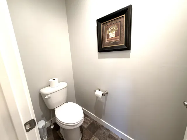 White toilet with a round bowl, light gray walls, dark patterned flooring, and a stainless steel toilet paper holder